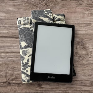 Pode incluir: Uma capa para leitor de e-books de tecido com padr&atilde;o preto e branco com um leitor de e-books Kindle preto dentro. O leitor de e-books tem uma tela branca e a palavra "kindle" impressa na parte inferior.