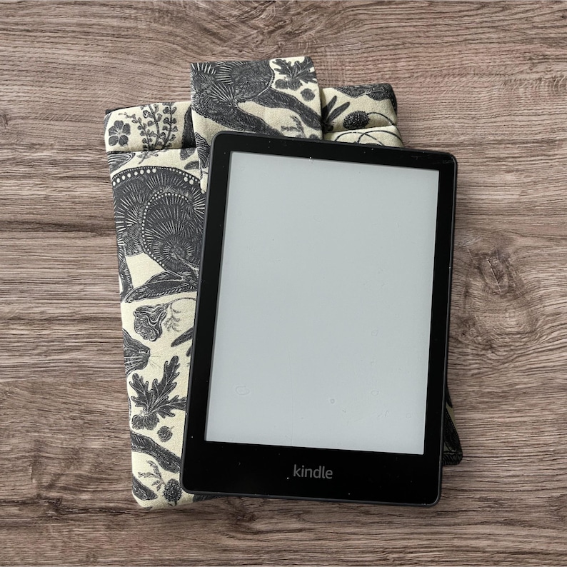 Pode incluir: Uma capa para leitor de e-books de tecido com padr&atilde;o preto e branco com um leitor de e-books Kindle preto dentro. O leitor de e-books tem uma tela branca e a palavra "kindle" impressa na parte inferior.