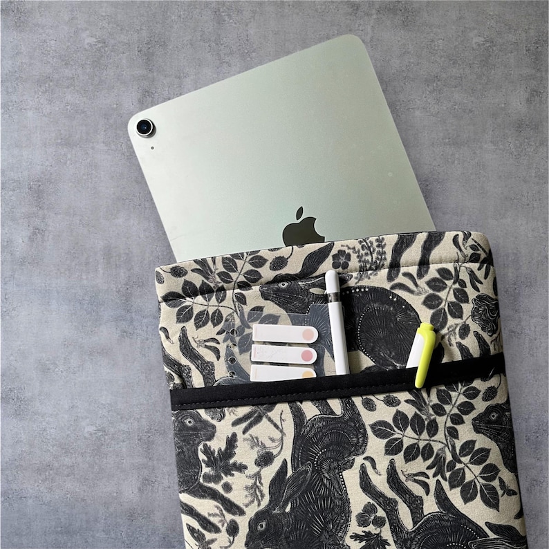 Pode incluir: Uma capa de tecido com padr&atilde;o preto e branco com um z&iacute;per preto e uma faixa el&aacute;stica preta. A capa cont&eacute;m um tablet prateado e uma caneta stylus preta. H&aacute; tamb&eacute;m tr&ecirc;s notas adesivas rosa e branco dentro da capa.