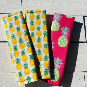 May include: Three fabric items with pineapple patterns. Two are white with yellow pineapples and green leaves. The third is pink with blue and green pineapples. The items are likely accessories or decorative pieces.