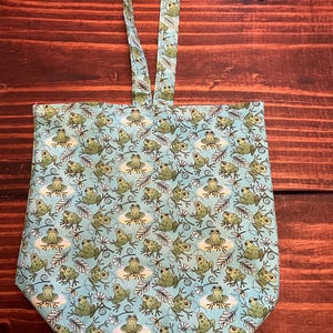 May include: A light blue fabric tote bag featuring a repeating pattern of green frogs, white flowers, and dark brown leafy sprigs. The bag has a single vertical handle attached to the top center, constructed from the same patterned fabric.