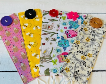 May include: Five fabric mask holders with various patterns and button closures. Designs include hedgehogs, bees, floral, owls, and a floral pattern with bees. Each holder has white elastic loops for attaching to a mask.