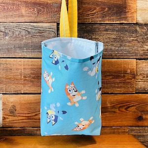 May include: A blue fabric bag with a yellow handle. The bag has a repeating pattern of cartoon dogs with orange, blue, and yellow colors. The bag is hanging on a wooden wall.
