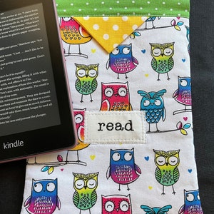 May include: A white fabric book cover with a colorful owl pattern. The cover has a green and yellow flap with white polka dots. The cover has the word "read" stitched on it in white thread.