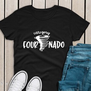 May include: Black t-shirt with a white graphic of a tornado and the text "category fournado". The shirt is laid flat on a wooden surface with a pair of white sneakers and blue jeans.