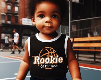 May include: A young child wearing a black basketball jersey with the text "Rookie of the Year" and a basketball graphic. The child is holding a small orange basketball and wearing black and white basketball shorts.