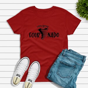 May include: Red t-shirt with a black graphic of a tornado and the text "category four NADO". The shirt is laying on a white wooden surface with a pair of white sneakers and blue jeans.