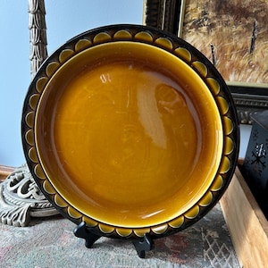 May include: A round, ceramic serving dish with a glossy, amber-colored center and a decorative black rim. The rim features a scalloped design. The dish is displayed on a black stand, showcasing its vintage aesthetic.