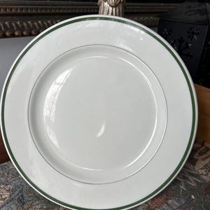 May include: White ceramic plate with a green border and a thin silver accent line. The plate has a simple, classic design, suitable for everyday use or special occasions. The plate is round.