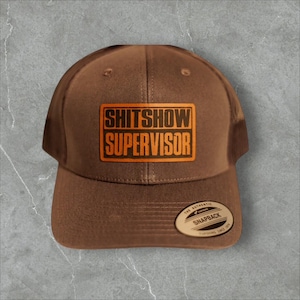 May include: Brown trucker hat with the text "SHITSHOW SUPERVISOR" in orange. The hat has a mesh back and a curved brim. A small, round tag is attached to the side.