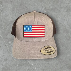 Può includere: Cappellino da camionista grigio con retro in rete marrone e toppa rettangolare con la bandiera americana. Il cappellino ha una visiera curva e una chiusura a scatto. L'etichetta recita "The Authentic Snapback Yupoong Since 1974."