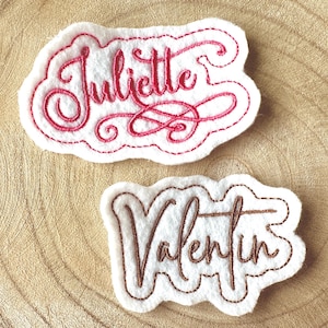 Peut inclure: Deux écussons de noms en feutre. L'un porte le nom "Juliette" brodé en script cursive rouge, souligné de coutures rouges. L'autre porte le nom "Valentin" brodé en script cursive marron, souligné de coutures marron.