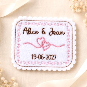 Peut inclure: Un écusson rectangulaire en feutrine blanche aux bords festonnés avec le texte brodé en marron Alice & Jean, un motif de deux cœurs brodés en rose et la date 19-06-2027 brodée en marron, entouré d'une bordure décorative rose.