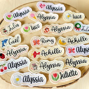 Peut inclure: Une collection d'étiquettes nominatives en tissu blanc aux bords festonnés, présentant des noms brodés tels qu'Alyssia, Jules, Luce, Gaël, Romy, Achille et Juliette, accompagnés d'icônes brodées colorées comme des fleurs et des animaux.