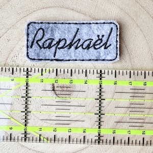 Peut inclure: Un écusson en feutre gris clair avec le nom "Raphaël" brodé en noir en écriture cursive. L'écusson est rectangulaire avec une bordure cousue noire. Une règle transparente avec des marques noires est en dessous.