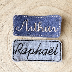 Peut inclure: Deux étiquettes de nom en feutre avec des noms brodés. L'étiquette du haut est bleue avec le nom "Arthur" brodé en fil beige. L'étiquette du bas est grise avec le nom "Raphaël" brodé en fil noir.
