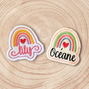 Peut inclure: Deux écussons brodés en forme d'arc-en-ciel. L'un porte le nom "Lily" en cursive, l'autre "Océane". Chaque écusson a un cœur rouge au centre de l'arc-en-ciel. Sur une surface en bois.