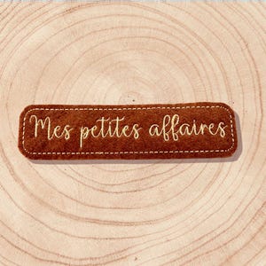 Peut inclure: Un patch rectangulaire en feutre marron avec le texte brodé "Mes petites affaires" en écriture cursive. Le patch est bordé d'une couture blanche pointillée. Le fond est une surface en bois clair avec des cercles concentriques.