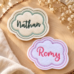 Peut inclure: Deux écussons en feutrine en forme de nuage avec des prénoms brodés. L'écusson du haut est crème avec une bordure bleu sarcelle et le prénom Nathan en vert foncé. L'écusson du bas est blanc avec une bordure rose et le prénom Romy en rose.