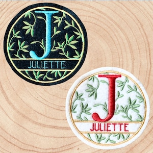 Peut inclure: Deux écussons brodés ronds. L'un présente la lettre "J" turquoise et le nom "JULIETTE" sur fond noir avec des motifs de feuilles vertes. L'autre a un "J" rouge et "JULIETTE" sur fond blanc avec des motifs de feuilles vertes.