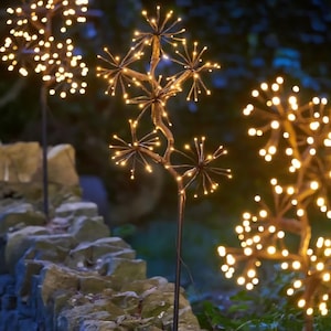 May include: Outdoor decorative lights shaped like trees with warm white LED lights. The lights are arranged on dark brown metal branches and stems. The lights are placed in a garden setting with a stone wall in the foreground.