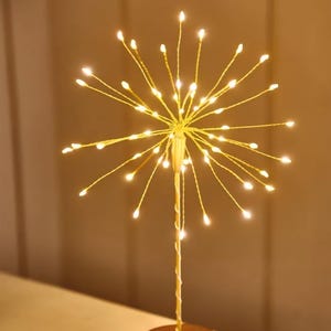 May include: A gold-coloured decorative light fixture with a starburst design. The fixture has a round base and a central stem from which numerous thin, wire-like arms extend, each tipped with a small, warm white LED light. The lights are illuminated, creating a soft glow.