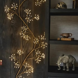 Può includere: Una lampada da parete decorativa a forma di ramo dorato con piccole luci bianche. Il ramo è montato su un pannello di legno scuro. Le luci, disposte a grappoli, creano un'atmosfera calda e accogliente.