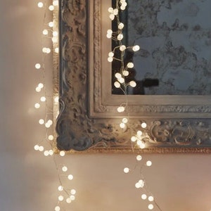 Op de afbeelding: Een string witte kerstverlichting gedrapeerd rond een decoratief spiegelframe. De lampjes zijn klein en rond en geven een warm licht.