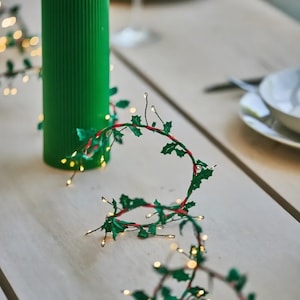 Puede incluir: Una vela verde acanalada con una llama encendida se encuentra sobre una superficie de madera clara. Una guirnalda de hojas verdes, tallos rojos y pequeñas luces blancas rodea la vela. La escena sugiere un tema festivo o navideño.