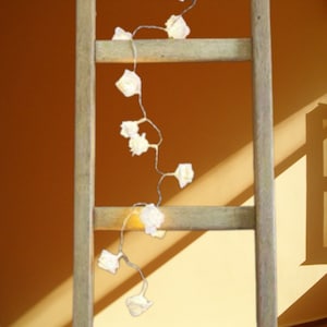May include: White string lights with small white flower-shaped bulbs are strung across a wooden ladder. The lights are hanging down from the top of the ladder and are illuminated.
