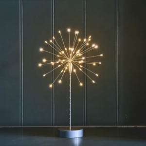 May include: A decorative table lamp with a starburst design. The lamp features numerous small, warm white LED lights radiating outwards from a central point. The base is a circular, gray metal. The lamp's stem is silver, and the background is a dark green.