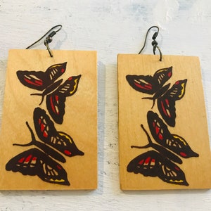 Può includere: Un paio di orecchini in legno con un design a farfalla. Le farfalle sono dipinte in nero, rosso e giallo su uno sfondo in legno marrone chiaro.