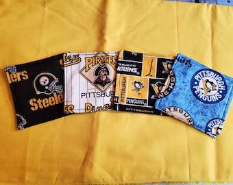 May include: Four fabric coasters featuring Pittsburgh sports team logos. The coasters display the Steelers, Pirates, and Penguins. The Steelers coaster is black with a gold helmet. The Pirates coaster has a baseball design. The Penguins coasters are black and blue.