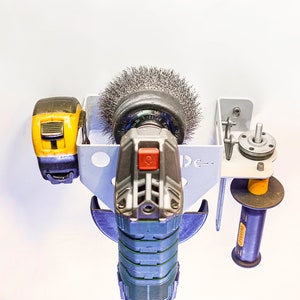 May include: A wall-mounted tool holder with a black and green angle grinder, a yellow and black power tool, and a black and yellow sanding disc. The holder is made of metal and has a white finish.