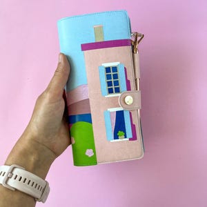 May include: A pink and blue house-shaped planner with a pen. The planner features a pink house design with blue window frames and a green lawn. The background is a blue sky with a pink and purple landscape.