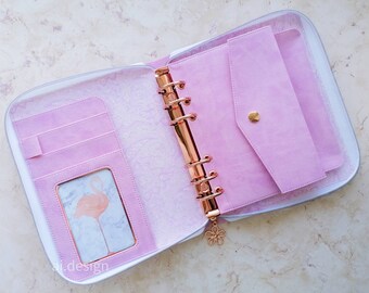 Puede incluir: Agenda rosa abierta con cremallera blanca, con una carpeta dorada rosa y un sobre rosa a juego. El interior incluye ranuras para tarjetas y un marco de fotos con una imagen de flamenco. Un dije de flor cuelga de la carpeta. El texto "ai.design" es visible.
