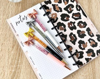 May include: A planner with a leopard print cover is open to a page with grid lines and the word "notes". Four pens with bow-shaped tops and glittery barrels in pink, silver, gold, and orange are on the page. Decorative items are in the background.
