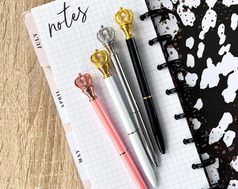 May include: An open notebook with a grid pattern and the word "notes" in cursive. Four pens with crown-shaped tops in gold, silver, and black are arranged on the notebook. The pens are pink, white, silver, and black.