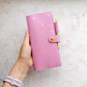 Könnte beinhalten: Ein rosa Planer mit goldenen Herzakzenten und einem goldenen Stifthalter. Der Planer wird mit einem goldenen Druckknopf verschlossen. Der Stift ist goldfarben mit einem Mickey Mouse-förmigen Aufsatz. Der Planer wird in einer Hand gehalten.