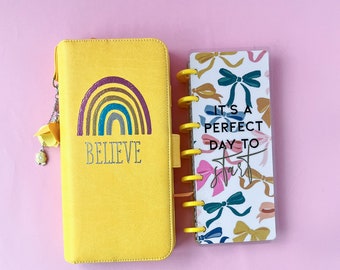 Può includere: Un'agenda gialla e un piccolo taccuino. L'agenda presenta un design arcobaleno e la parola "BELIEVE". La copertina del taccuino dice "IT'S A PERFECT DAY TO START" e ha un motivo a fiocco. Entrambi gli articoli hanno dettagli gialli.