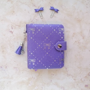 May include: A purple planner with a bow and dot pattern, silver accents, and a tassel charm. Two purple bow paperclips are also present. The planner has a button closure. The text "ai.design" is visible.