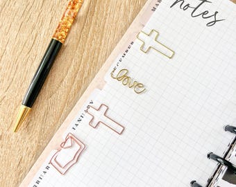 以下が含まれることがあります： 金色のリボン付きペン、黒い軸と金色のアクセントが、木製の表面にプランナーの隣に置かれています。 プランナーには、十字架の形をした金と銅色のペーパークリップと「love」の文字があります。