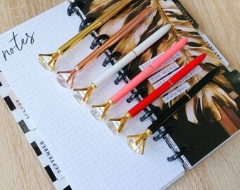 May include: A collection of six pens with diamond-shaped tops in gold, rose gold, white, pink, red, and black. The pens rest on a notebook with grid paper and monthly dividers. The word "notes" is visible.