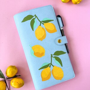 May include: A light blue planner with a lemon design. The planner has a gold snap closure and a pen holder with a black pen. Yellow lemons and green leaves are featured on the cover. The background is pink.