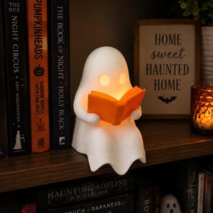May include: A white ghost-shaped lamp with a smooth matte finish, featuring two hollow circular eyes that emit a warm orange glow. The ghost is holding an open, bright orange book in its hands, which is also illuminated from within the figure.