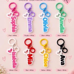 May include: Eight custom name keychains featuring a vertical name tag, a heart-shaped charm, and a circular ring with a lobster clasp. The names Sophia, Emma, Olivia, Amelia, Isabella, Mia, Chloe, and Ava are displayed in various colors including pink, purple, blue, mint, white, red, yellow, and black.