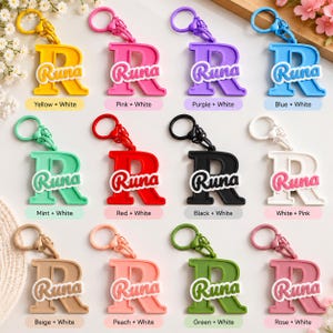 May include: A collection of twelve letter R keychains in various colors, each featuring the name Runa in a contrasting script font. The keychains include yellow, pink, purple, blue, mint, red, black, white, beige, peach, green, and rose colors.