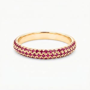 Puede incluir: Un anillo de banda de oro con un engaste pavé de numerosos rubíes pequeños, redondos y de color rojo rosado intenso que rodean toda la superficie exterior de la banda de metal, creando una apariencia texturizada continua de gemas en oro amarillo.
