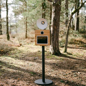 May include: A wooden photo booth with a white flash and a black screen, mounted on a black metal stand. The photo booth is in a forest setting.