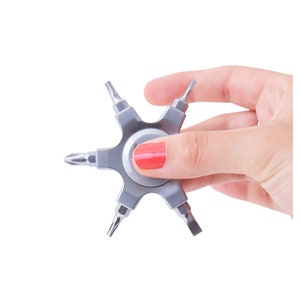 May include: A silver multi-bit screwdriver tool with six different screwdriver heads. The tool is held in a hand with red nail polish. The tool is designed to be compact and versatile for various repair tasks.